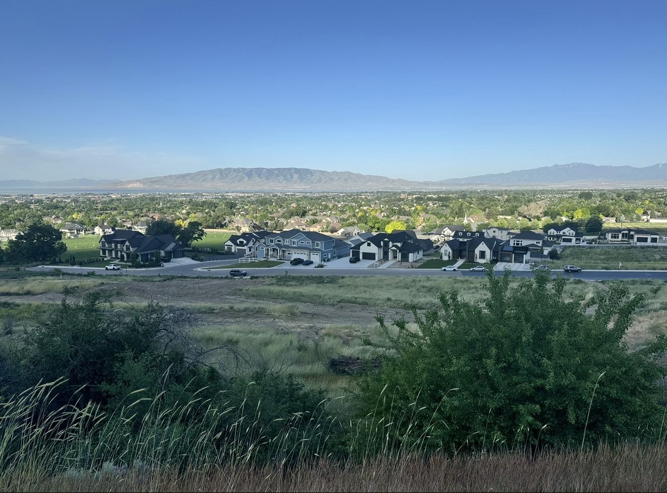 Elevated view from the trail over grassy foothills and a row of houses, with tree-filled Utah Valley, a sliver of Utah Lake, and mountains on the horizon.